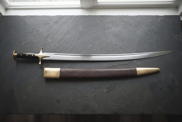 Hand forged Indian sword of talwar type with a mirror polished curved blade and brass disc pommel, laid on dark slate beside its brass mounted leather scabbard