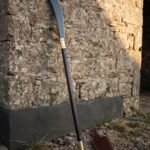 Medieval glaive with a long blackened curved steel blade and brass langets on a dark haft leaning against a rough limestone barn wall on gravel