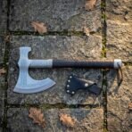 Overhead view of the norse runic axe on grey flagstone paving beside its black leather head sheath, showing the full crescent bit and rune engraved steel.