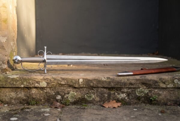 Hand forged damascus side sword with a broad layered leaf shaped blade and polished four ring hilt on a sandstone step beside its russet leather scabbard
