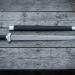 Hand forged Damascus Chinese sword with a straight layered double edged jian blade and wrapped grip lying on weathered timber decking beside a black scabbard