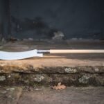 Hand forged glaive weapon with a long polished curved steel blade on a pale ash haft resting on a lichened stone step in a doorway