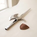 Polished steel halberd head with an axial spike, crescent blade and open socket lying on a pale surface beside a brown fitted leather blade cover