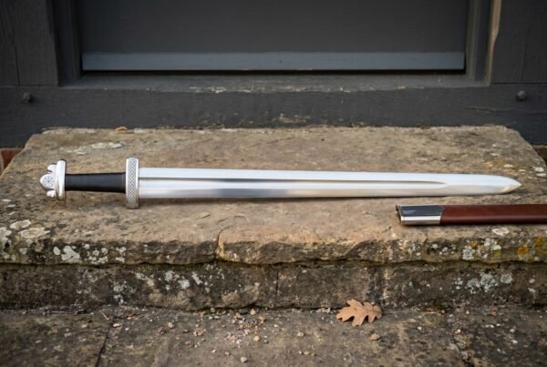 Hand forged Carolingian sword with polished fullered blade, long straight crossguard and brown leather scabbard resting on a weathered stone doorstep