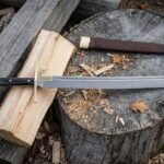 Hand forged Damascus messer with a layered patterned straight backed blade and brass fitted hardwood grip resting on a split log and tree stump beside a leather sheath