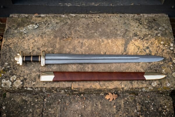 Hand forged Viking sword with a bright polished high carbon steel blade and brown leather scabbard laid across a warm sandstone ledge in daylight
