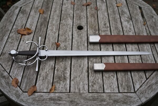 Hand forged side sword with a broad fullered blade and polished swept hilt lying on a weathered timber garden table beside its brown leather scabbard