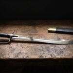 Hand forged talwar sword with a mirror polished curved blade, blackwood grip and disc pommel on a scarred dark timber workbench beside its black leather scabbard