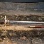 Hand forged dueling sword with a gilt brass swept hilt and rosewood grip resting on a sunlit stone step beside its russet leather scabbard with brass throat