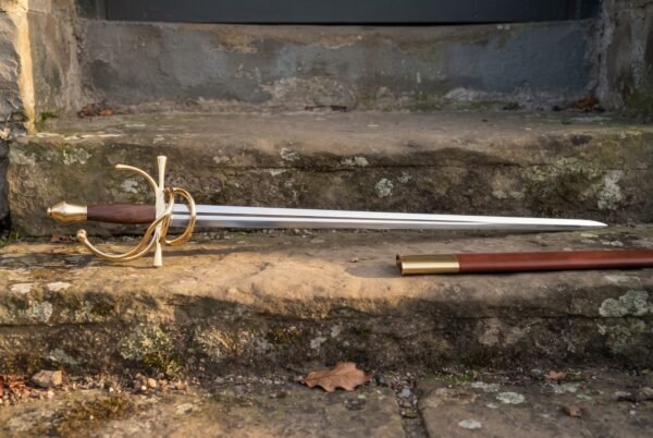 Hand forged dueling sword with a gilt brass swept hilt and rosewood grip resting on a sunlit stone step beside its russet leather scabbard with brass throat