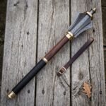 Damascus mace with six forge welded flanges, brass collars and a rosewood haft lying on a silvered timber table beside a buckled leather wrist strap