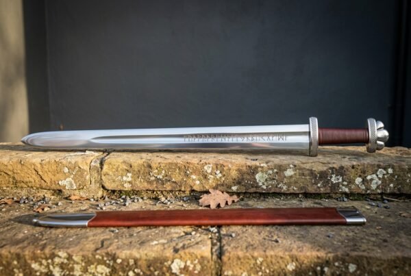 Hand forged viking rune sword with a broad blade carrying a darkened Elder Futhark inscription, red leather grip and brown scabbard on a sandstone ledge