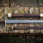 Hand forged Damascus arming sword with bone disc pommel beside a rune tooled leather scabbard and matching belt, laid out on a lichen covered brick ledge.