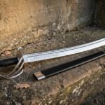 Hand forged high carbon steel cavalry sabre with a three bar steel guard, ribbed black leather grip and rune engraved curved blade resting on a weathered sandstone step beside its black leather scabbard