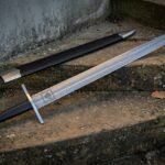 Hand forged Damascus bastard sword with an etched Valknut panel and long black leather grip, lying with its black scabbard on weathered stone steps.