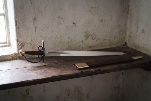 Hand forged espada ropera with a broad polished blade and blackened brass capped hilt on a dark oak shelf beside its brass mounted leather scabbard