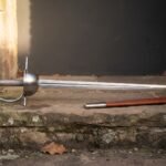 Hand forged spanish rapier with a layered damascus blade and deep polished steel cup hilt resting on a sunlit stone step beside its tan leather scabbard