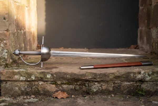 Hand forged spanish rapier with a layered damascus blade and deep polished steel cup hilt resting on a sunlit stone step beside its tan leather scabbard