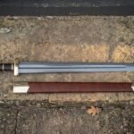 Hand forged Viking sword with scabbard in brown leather and a lobed steel pommel, laid on a sandstone step beside its belt in daylight
