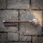 Hand forged gothic mace with a polished steel spiked morning star head and walnut haft lying on grey flagstone paving beside a leather wrist strap