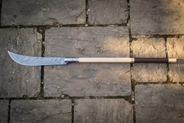 Damascus glaive with a ladder patterned forge welded blade and pale ash haft with a leather wrapped grip lying on grey cobbled flagstones