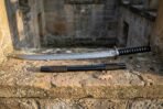 Hand forged wakizashi with black cord tsuka on a sandstone ledge in an old stone doorway, black saya with brown cord wrap set below the polished blade