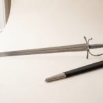 Hand forged renaissance short sword with a broad clipped point blade and polished knuckle bow hilt on a pale studio backdrop beside its black leather scabbard