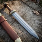 Hand forged Damascus gladius with a raindrop pattern Pompeii blade, ribbed walnut grip and brass guard, resting on a cracked tree stump beside its brown leather scabbard.