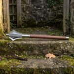 Hand forged flanged mace with six polished high carbon steel flanges and an American walnut haft resting on a mossy stone step beside a leather wrist strap