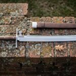 Hand forged messer sword with a polished straight backed blade and cream bone toned grip lying on a lichened red brick ledge beside a brown leather sheath