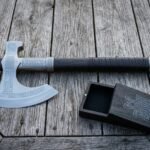 Viking axe gift set on a silvered timber table, the hand forged runic bearded axe laid beside its open rune carved wooden presentation box with linen lining.