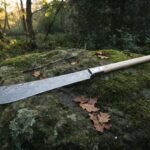 Damascus polearm with a long random pattern forge welded blade and pale ash haft resting on a moss covered boulder in autumn woodland