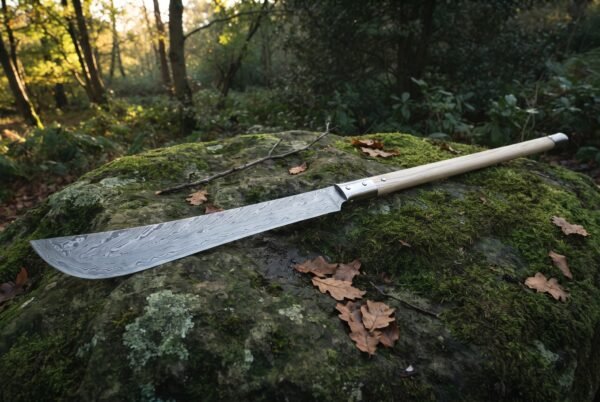Damascus polearm with a long random pattern forge welded blade and pale ash haft resting on a moss covered boulder in autumn woodland