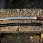 Hand forged high carbon steel katana with a deep crimson cord wrapped grip and black lacquered saya laid on a weathered stone bench scattered with fallen autumn leaves