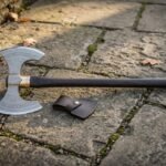 Long hafted hand forged viking war axe with an engraved steel head and pale ash handle, laid flat across grey flagstone paving in natural light