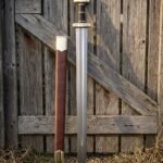 Hand forged knight sword replica in spring steel with a leather wrapped grip and brown scabbard leaning against a weathered timber farm gate