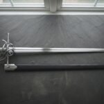 Hand forged swept hilt rapier with a coiled polished steel guard and wire wrapped grip laid on a dark slate slab beside its black leather scabbard