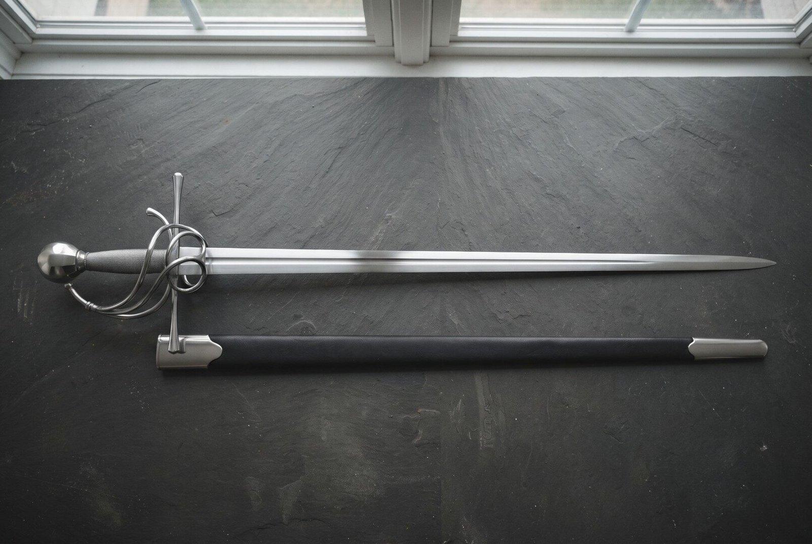 Gemini_Generated_Image_nnqfkjnnqfkjnnqf Hand forged swept hilt rapier with a coiled polished steel guard and wire wrapped grip laid on a dark slate slab beside its black leather scabbard