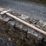 Hand forged polehammer with a polished steel hammer face, curved beak and top spike on a pale ash haft resting along a low drystone wall