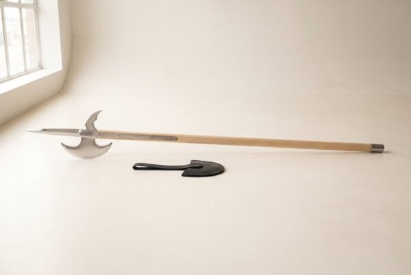 Medieval halberd laid flat on a pale barn floor, satin steel spike and crescent blade on a long ash haft with steel langets beside a black leather head cover
