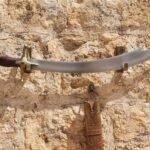 Hand forged Arabian sword with a curved polished steel blade and rosewood grip mounted on a sandstone fortress wall above its tooled leather scabbard
