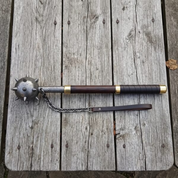 Damascus flail with a forge welded spiked ball head, brass collars, rosewood haft and hanging chain laid on a silvered timber table top