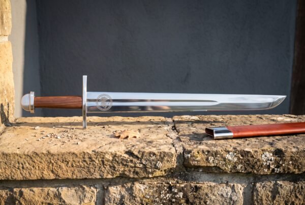 Hand forged medieval falchion with a broad straight backed polished blade and rosewood grip lying on a sandstone ledge beside a red leather scabbard