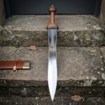 Hand forged Mainz gladius with a polished waisted leaf blade and ribbed cherry wood grip, standing upright on a lichened stone doorstep beside its tooled leather scabbard.