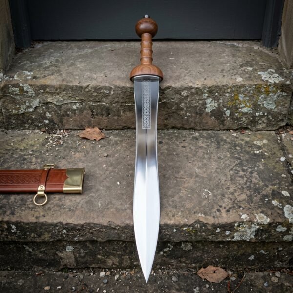 Hand forged Mainz gladius with a polished waisted leaf blade and ribbed cherry wood grip, standing upright on a lichened stone doorstep beside its tooled leather scabbard.