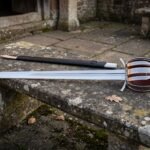 Hand forged basket hilt sword with a knotwork engraved steel shell guard and oxblood leather grip lying on a weathered stone garden bench beside its dark scabbard