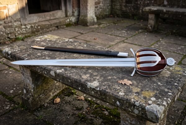 Hand forged basket hilt sword with a knotwork engraved steel shell guard and oxblood leather grip lying on a weathered stone garden bench beside its dark scabbard