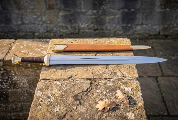 Hand forged ranger sword in high carbon steel with a leather wrapped grip and tan leather scabbard resting on a lichen covered stone slab outdoors