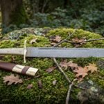 Hand forged Damascus seax with a layered single edged blade and pale polished bone handle lying on a moss covered boulder beside its brown leather sheath
