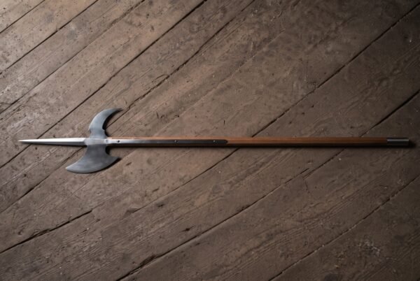 Battle halberd with a satin steel spike, crescent blade and hooked fluke on a long hardwood haft laid across a dark weathered plank floor